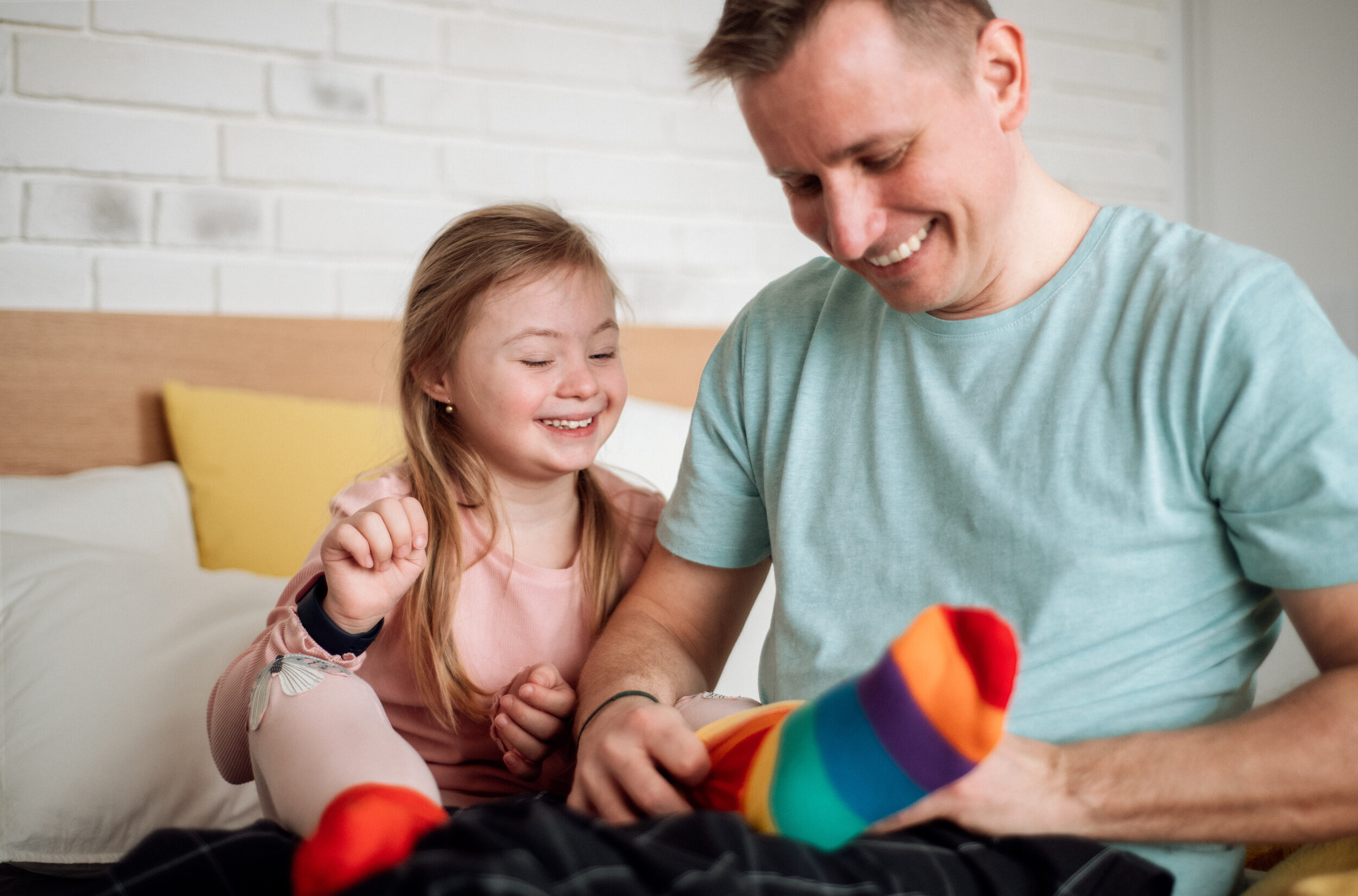 Father putting on different socks to his little daughter with Down syndrome when sitting on bed at home.